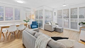 A modern living room with a dining area, a blue armchair, a grey sofa, and a ceiling fan. - Ocean and Pool Views from Beachside Apartment! (Gold Coast)