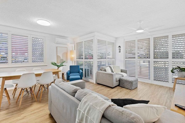 A modern living room with a dining area, a blue armchair, a grey sofa, and a ceiling fan. - Ocean and Pool Views from Beachside Apartment! (Gold Coast)