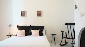 A bedroom with a bed, two pillows, a nightstand, a lamp, a rocking chair, and two framed pictures on the wall. - Chevignon country charm in the heart of town (Queenscliff)