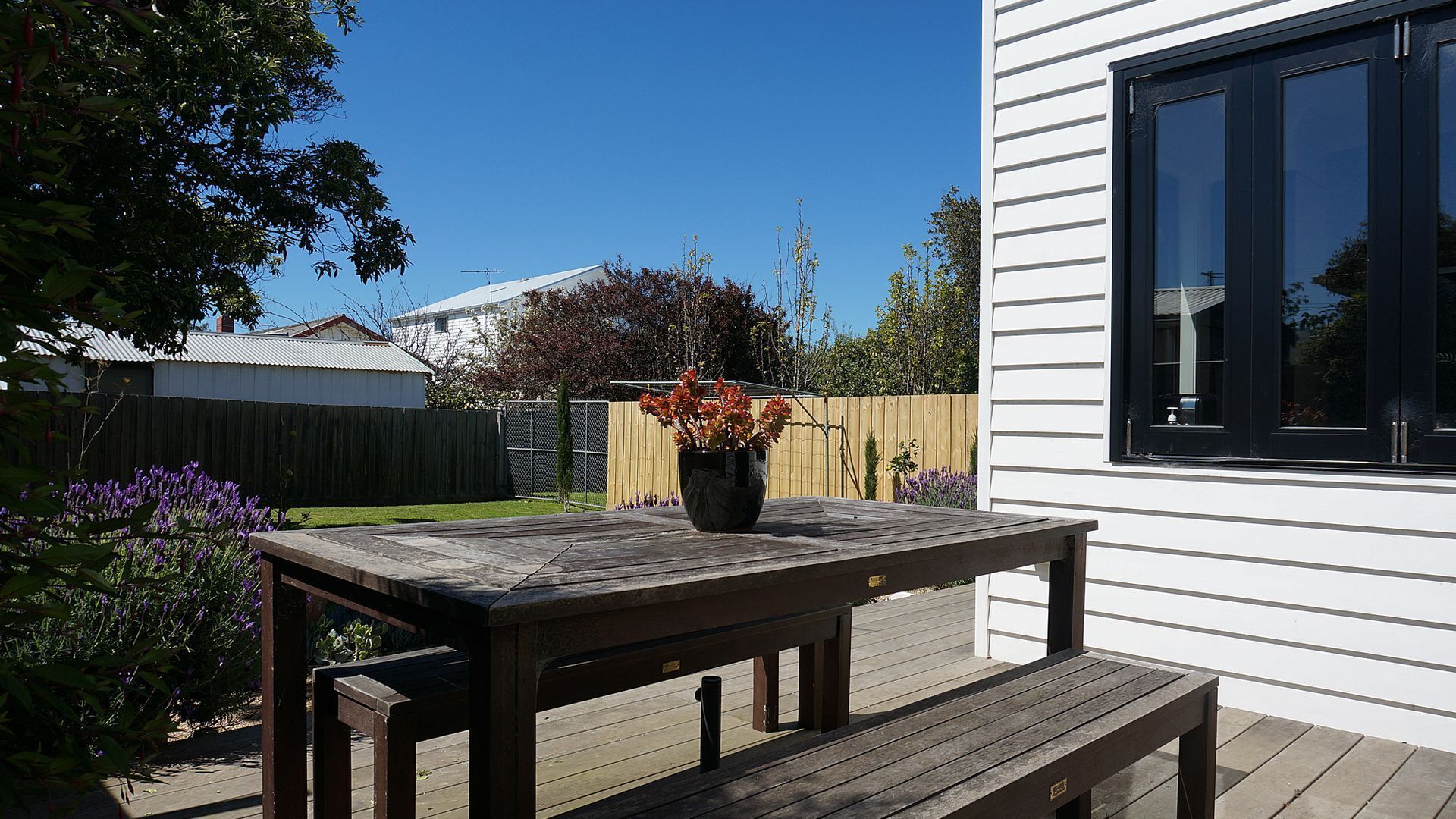 A wooden deck with a bench and a table, a potted plant with red flowers, and a white building with black-framed windows.