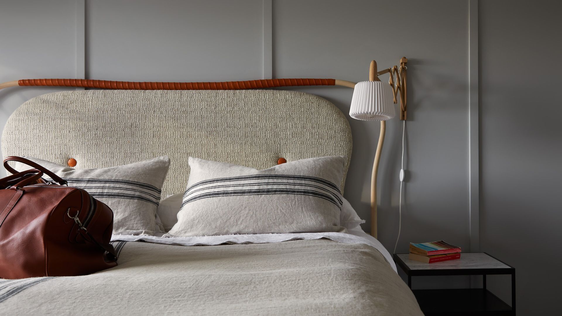 A bed with a headboard, pillows, a handbag, a bedside table with books, and a lamp.