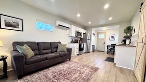 A modern living room with a dark brown sofa, a patterned rug, and a kitchen area with white cabinets, a refrigerator, and a microwave.