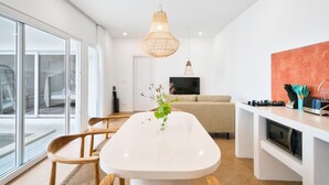 Een moderne keuken met een wit aanrecht, een eettafel met stoelen, een hanglamp en een televisie boven een bank.