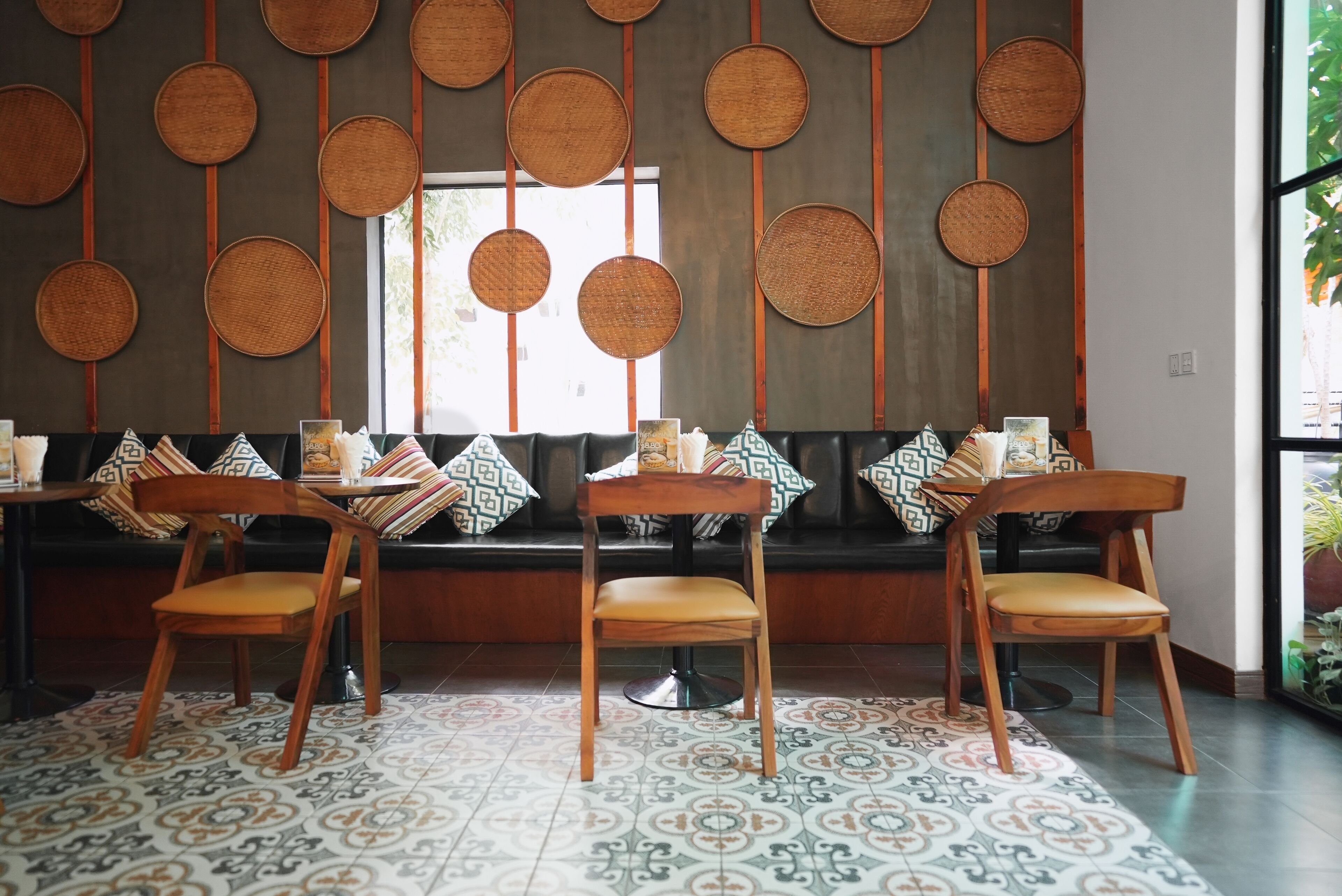 L’intérieur d’un restaurant présente une longue banquette, des chaises en bois, des carreaux de sol à motifs et un mur décoré de panneaux tissés de forme circulaire.