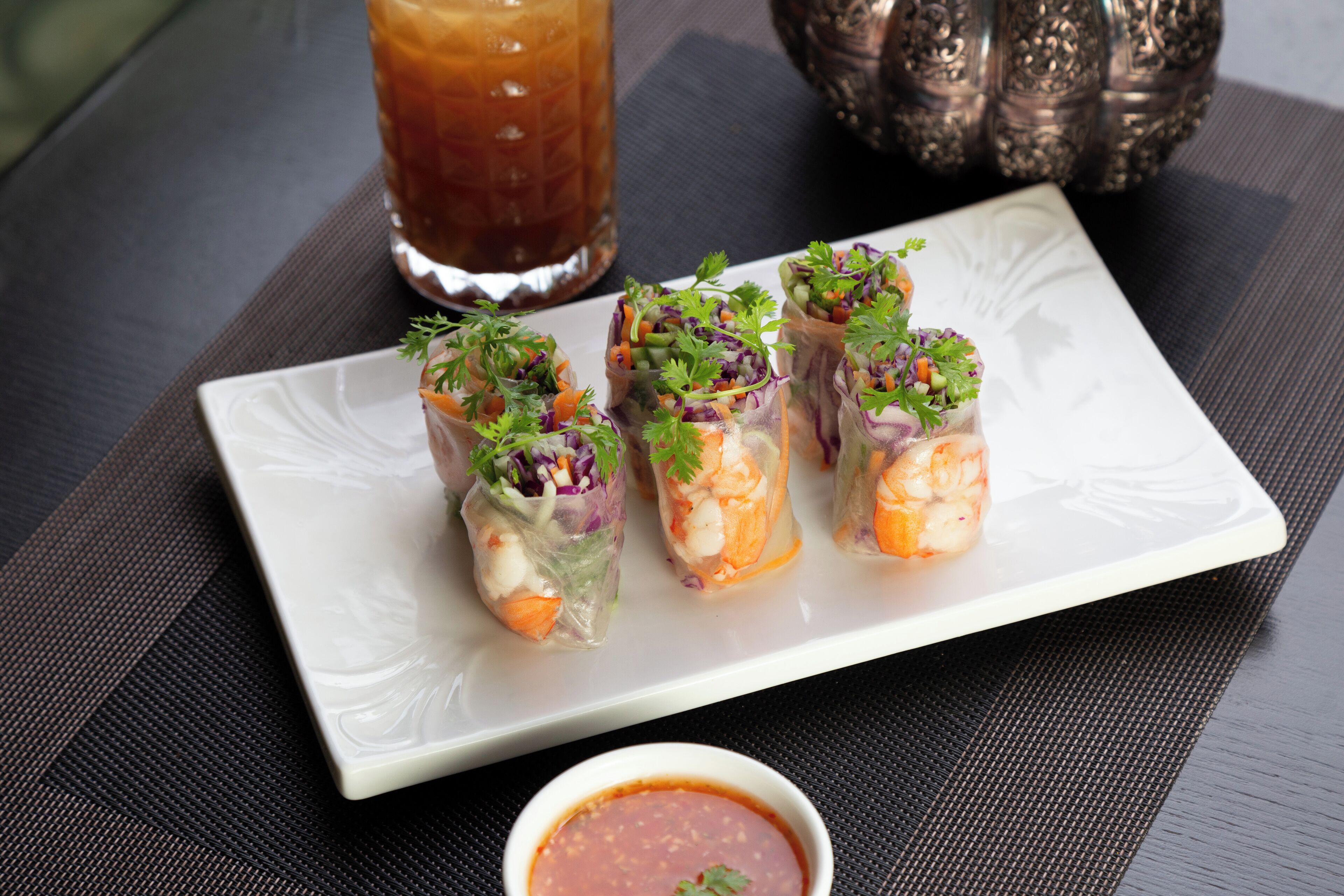 A white plate with four spring rolls garnished with herbs, a glass of iced tea, and a small bowl of sauce.