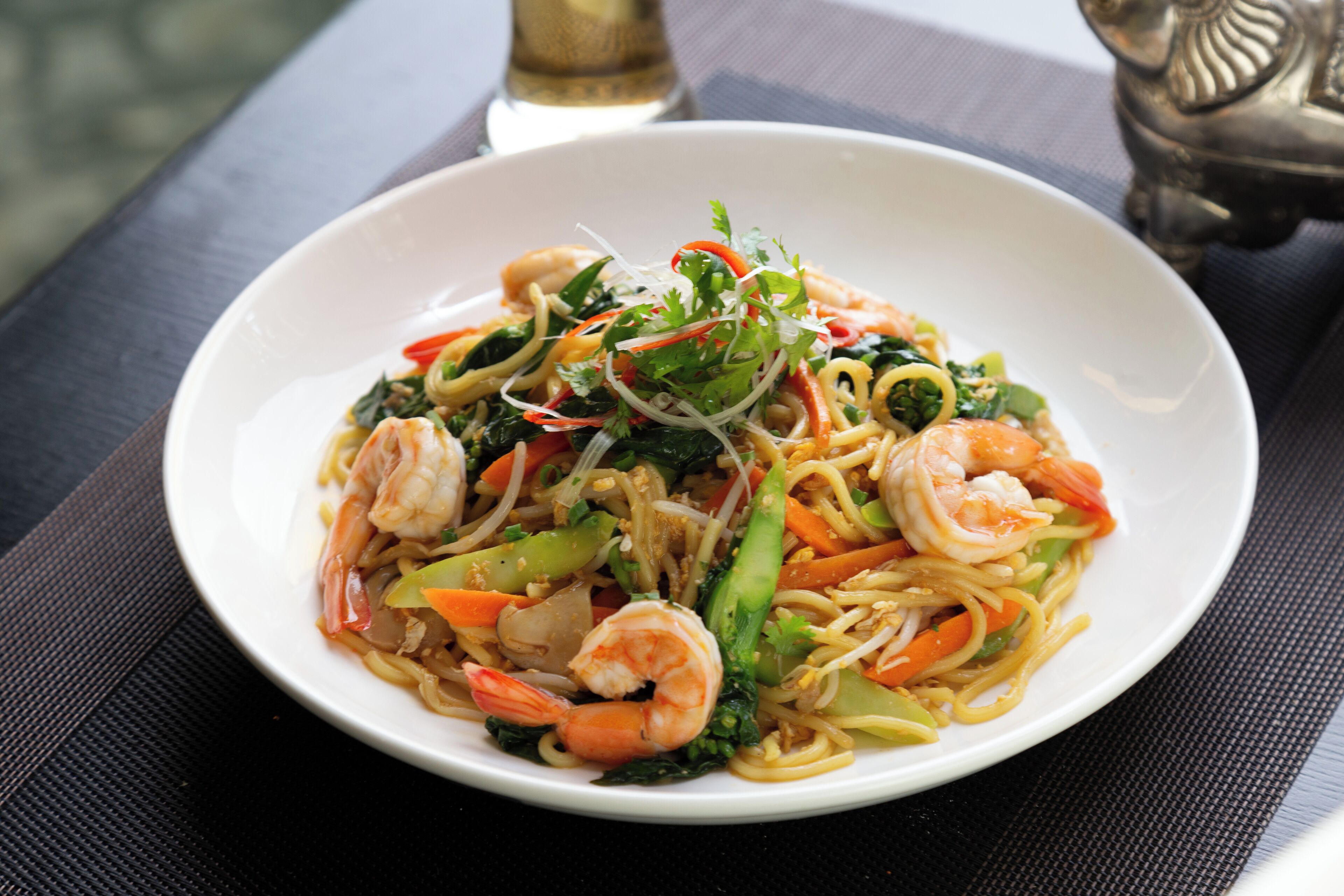 A bowl of stir-fried noodles with shrimp and vegetables on a dark table.