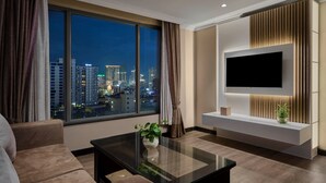 A modern living room with a large window, a flat-screen TV mounted on a wall, a sofa, a coffee table, and a potted plant. - Caravan Hotel by EHM (Phnom Penh)