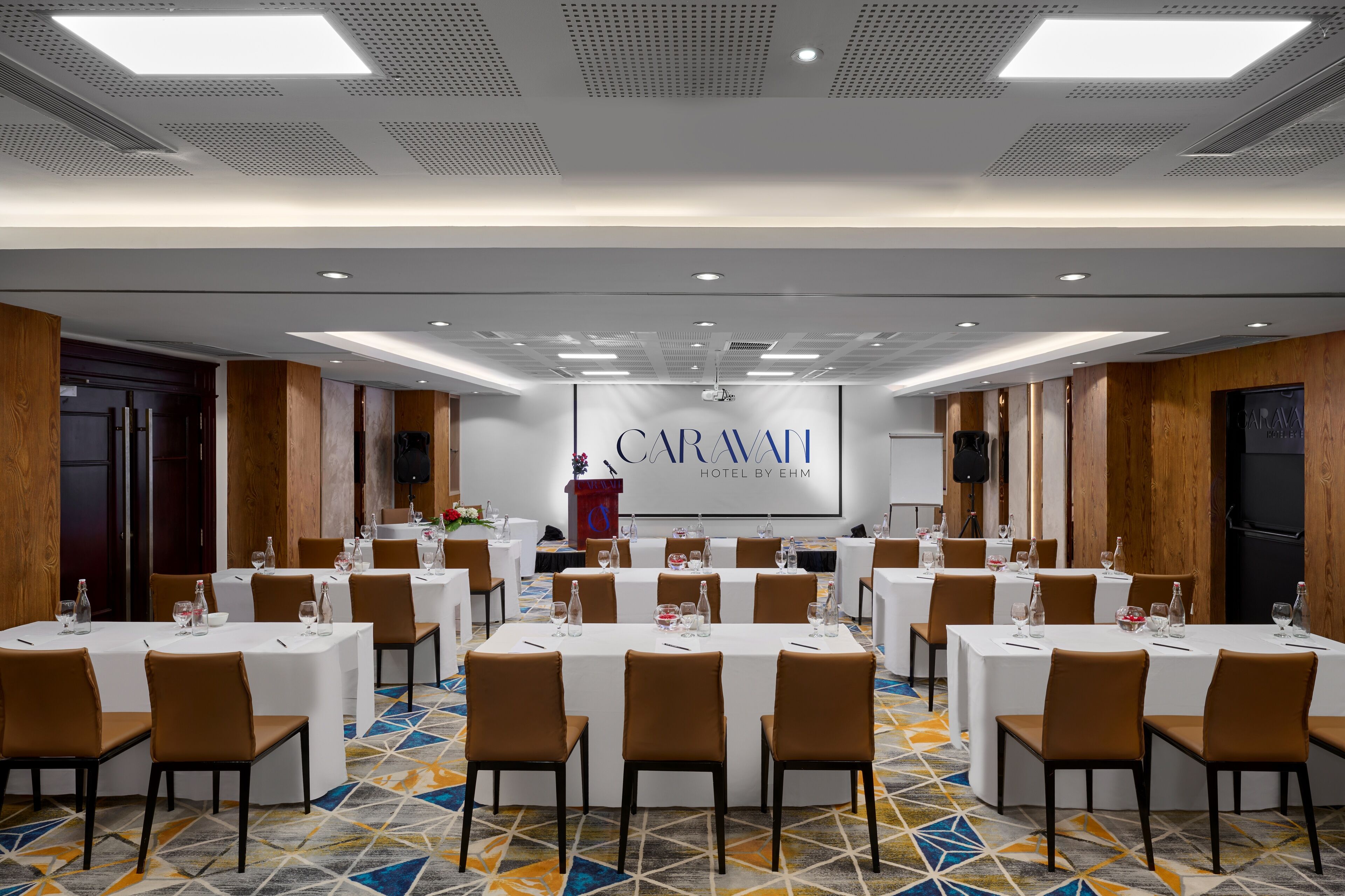 A conference room with multiple tables set for meetings, a podium, and a banner for Caravan Hotel by EHM.