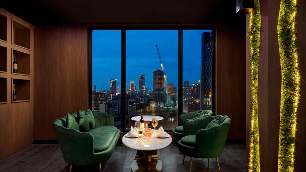 A modern room with a city view, a round table with a bottle and glasses, and green upholstered chairs. - Caravan Hotel by EHM (Phnom Penh)