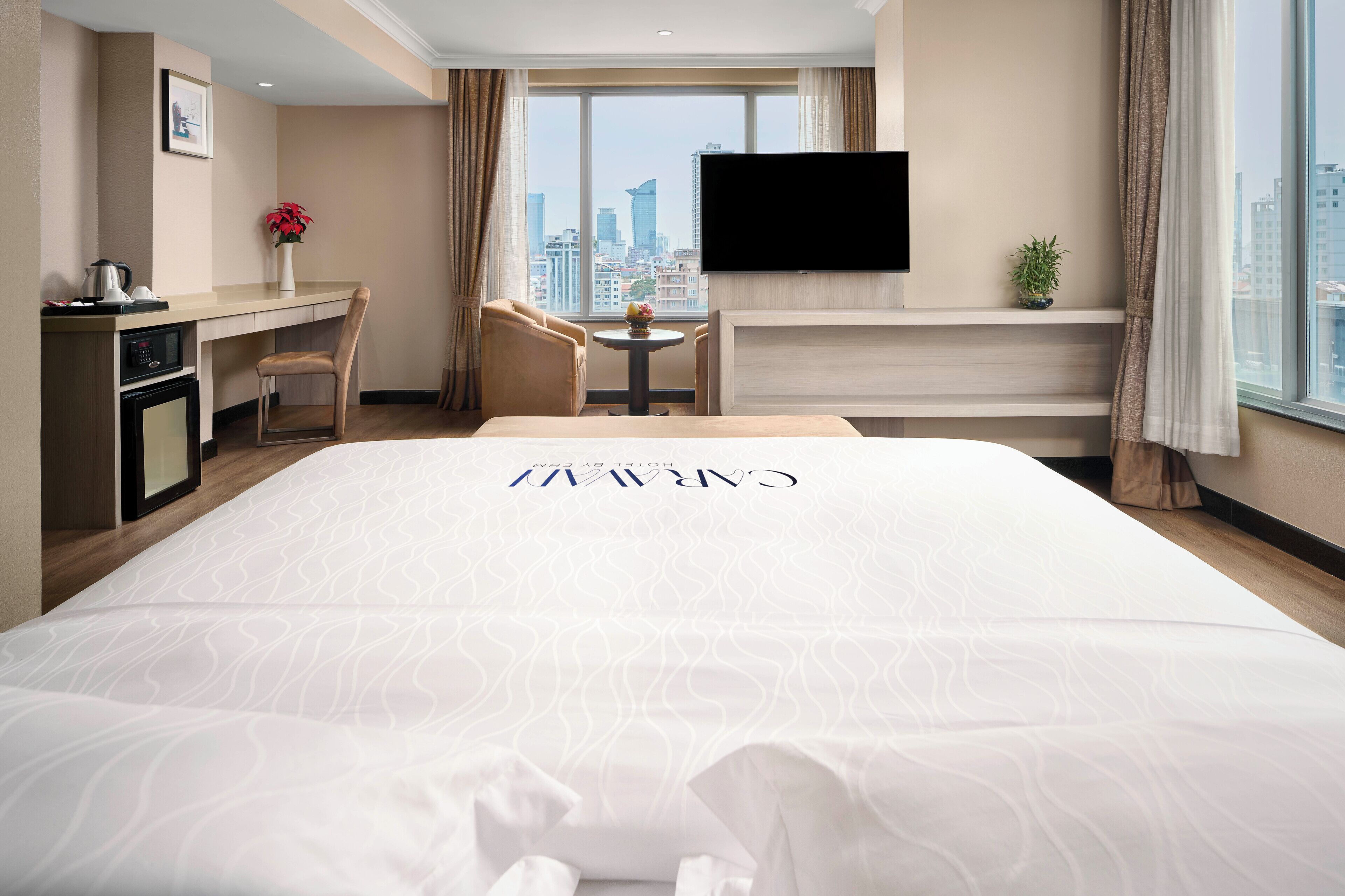 A hotel room with a large bed, a desk with a computer, a chair, a small table with a vase, a TV on the wall, and a city view.