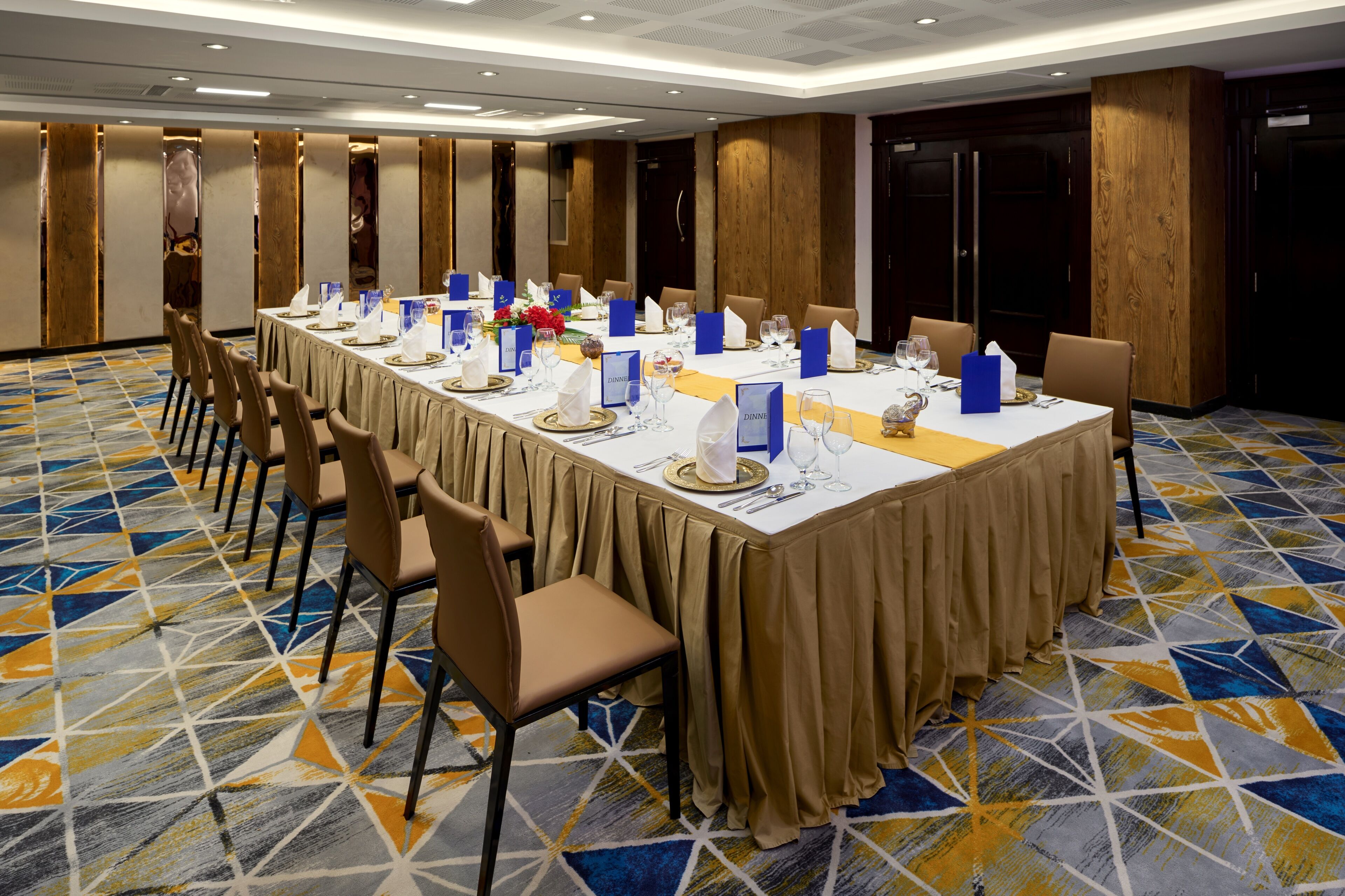 Un salón de conferencias con una larga mesa dispuesta para una reunión, rodeada de sillas, una alfombra estampada y paneles de madera.