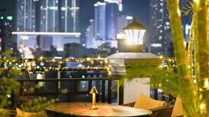 An outdoor dining area with a table and chairs, surrounded by greenery and string lights. In the background, there are illuminated high-rise buildings. - Caravan Hotel by EHM (Phnom Penh)