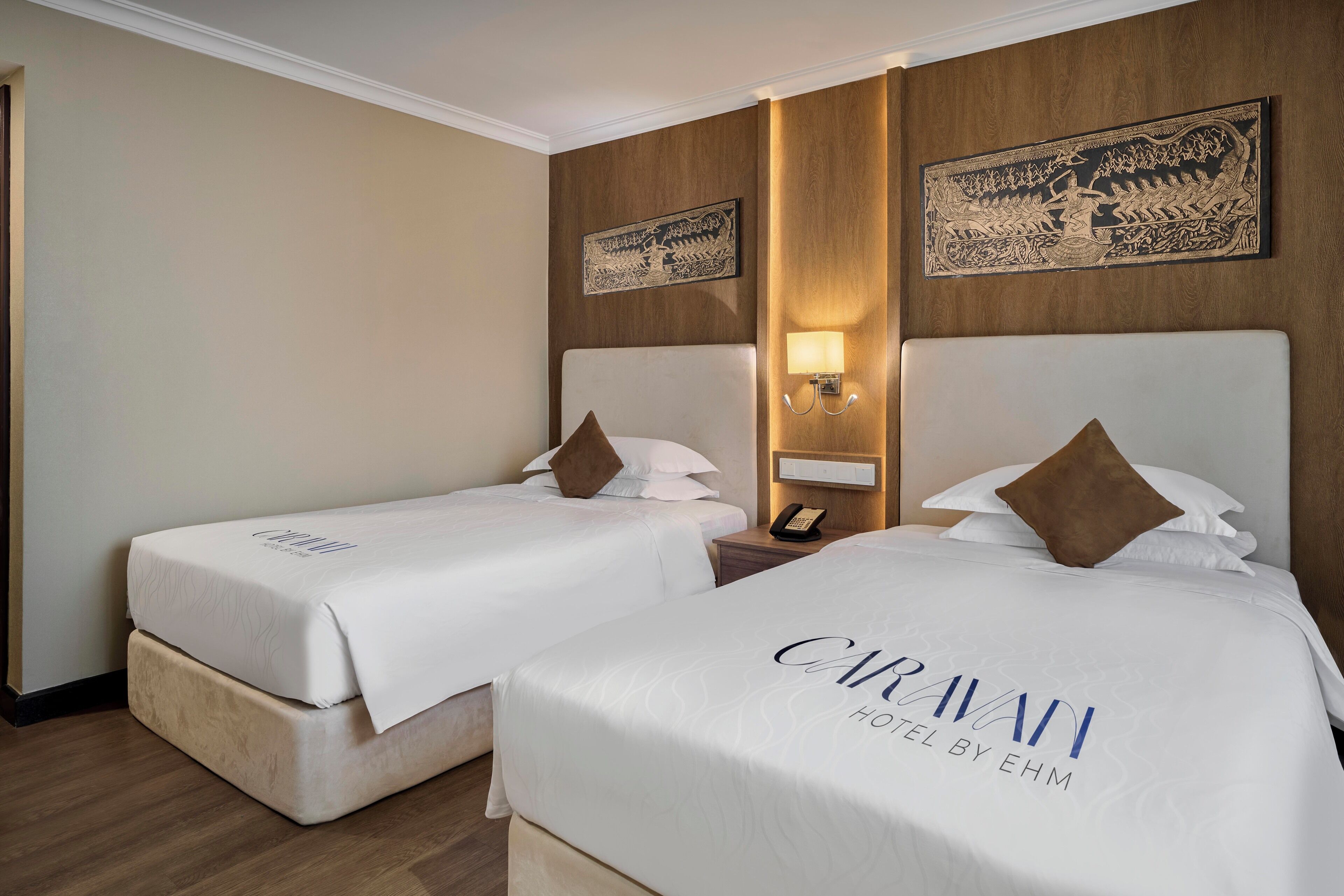 Two twin beds in a hotel room with a wooden headboard, a nightstand with a lamp, and artwork on the wall.