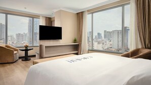 A hotel room with a large bed, a flat-screen TV mounted on the wall, a small table with a plant, and a view of the city through large windows. - Caravan Hotel by EHM (Phnom Penh)