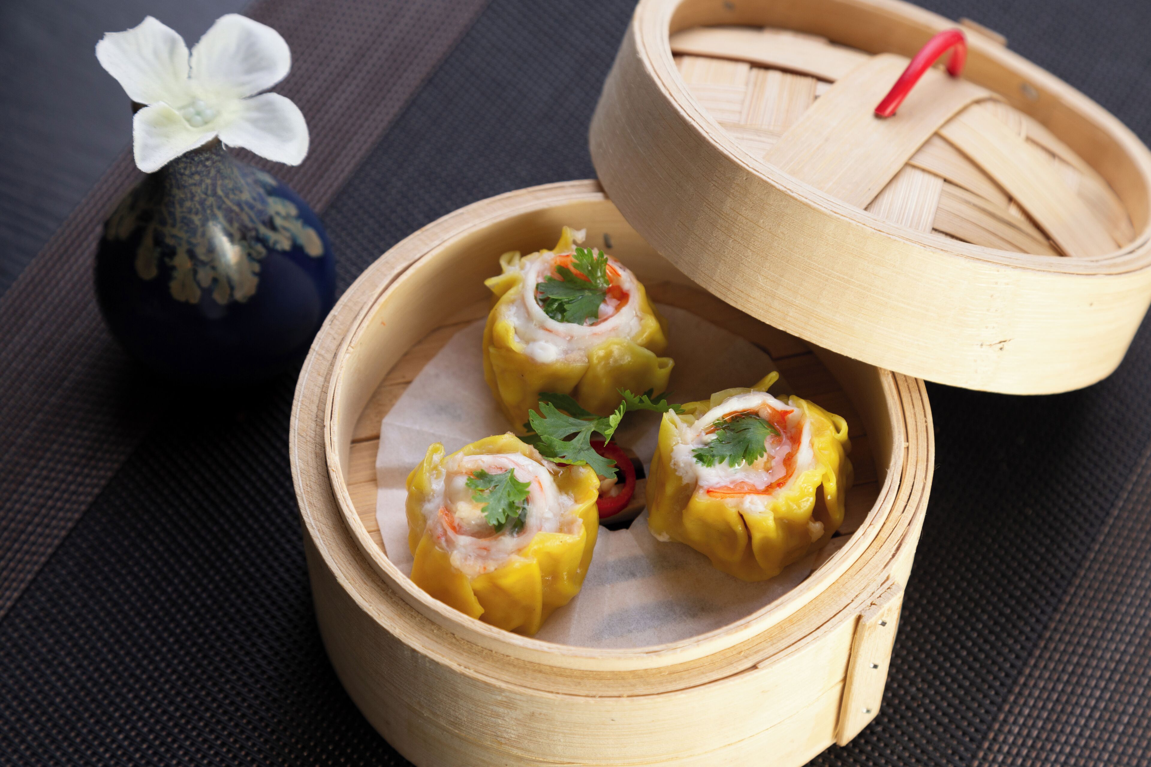 Una vaporera de bambú con tres dumplings, un jarrón pequeño con una flor y un palillo rojo.
