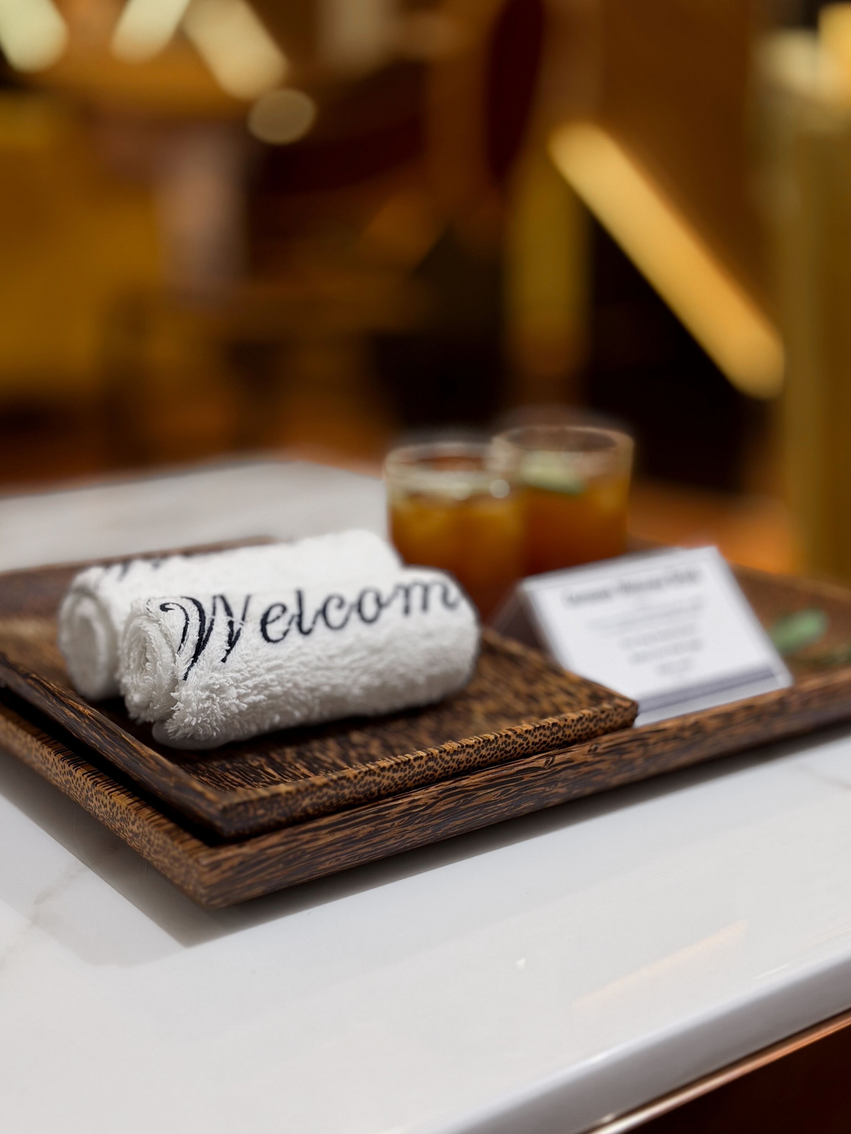 Ein gerolltes Handtuch mit der Aufschrift "Welcome", ein Glas Eistee und eine kleine Karte auf einer Holzschale.