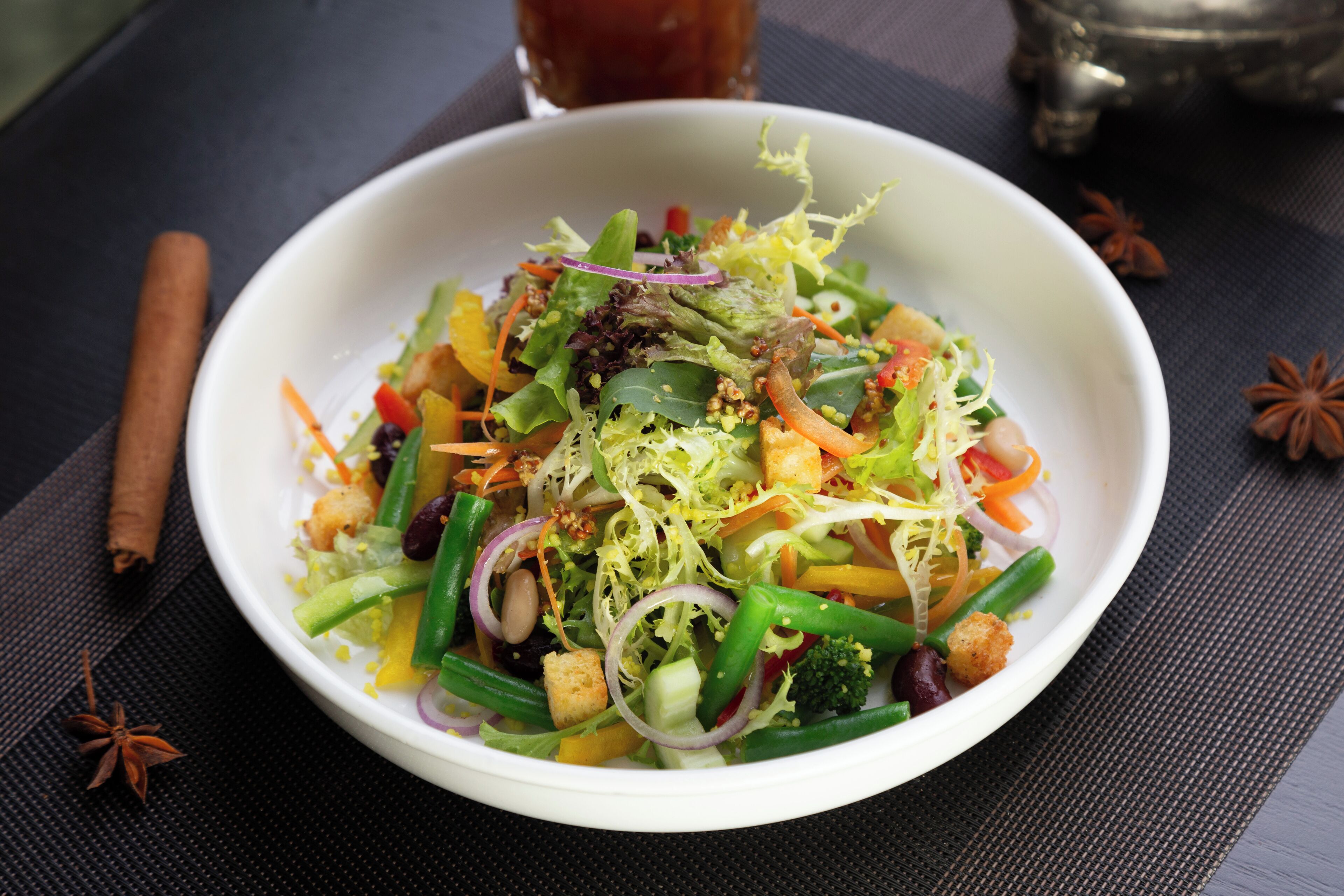 A bowl of colorful salad with various vegetables and nuts, placed on a dark table with a glass of tea and cinnamon sticks nearby.