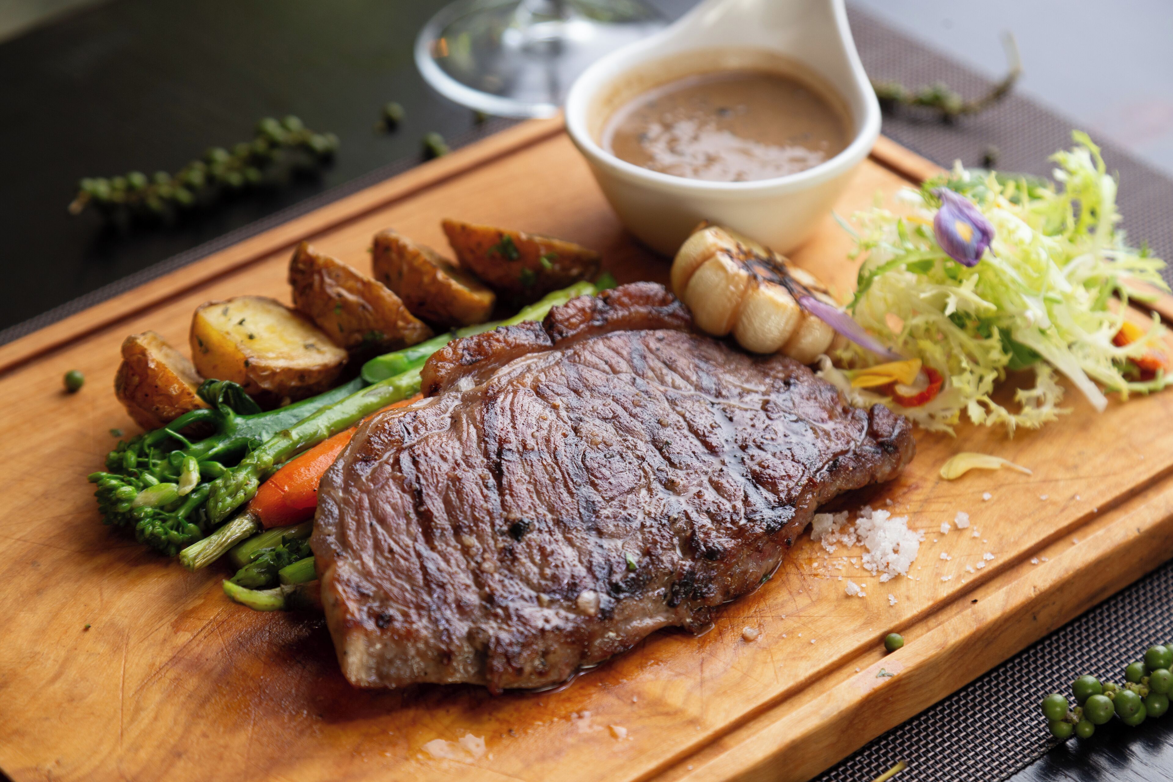 A steak with vegetables and a gravy boat on a wooden board.