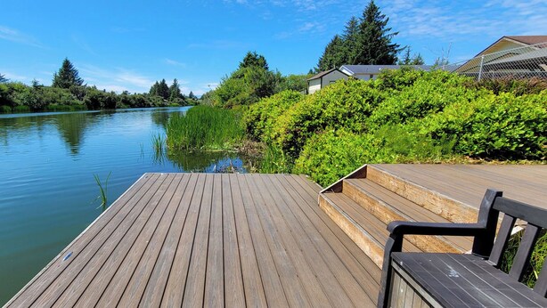 Une terrasse en bois avec un banc donnant sur une rivière paisible, entourée d’une végétation luxuriante et avec une maison en arrière-plan.