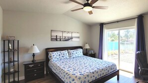 Une chambre à coucher comprenant un lit, une table de chevet, une lampe, un bureau, une chaise, un ventilateur de plafond, une fenêtre avec des rideaux et un téléviseur fixé au mur.