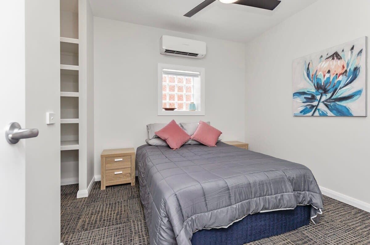 A bedroom with a bed, a window, an air conditioning unit, a painting, and a wardrobe.