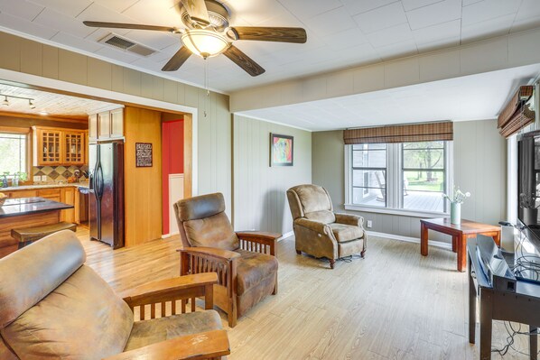 A spacious living area with a ceiling fan, wooden furniture, and a kitchen in the background.