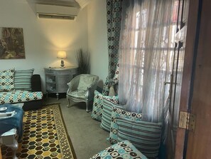 Ein Zimmer mit einem gemusterten Teppich, einer Sofagarnitur, einem Sessel, einer Lampe und einem Vorhang.