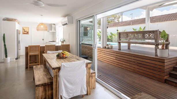 A modern kitchen with a wooden table and chairs, a ceiling fan, and a view of a wooden deck with a bench. - Pool - Ohana - Oceanstays (Yamba)