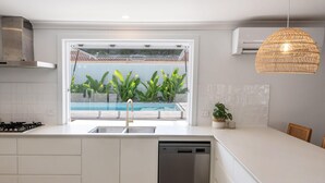 A modern kitchen with a large window, white cabinetry, a dishwasher, and a hanging light fixture. - Pool - Ohana - Oceanstays (Yamba)