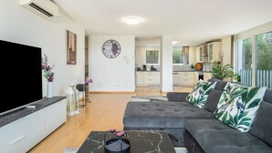 Una sala de estar moderna con un televisor de pantalla plana, un sofá gris con almohadones tropicales, una mesa de centro de mármol y una cocina al fondo.