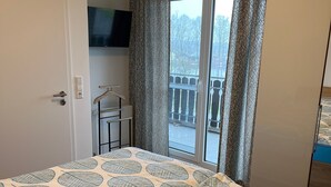 A hotel room with a bed, a TV mounted on the wall, a balcony with patterned curtains, and a view of the outside. - Seeblick Holiday Apartment   – Ideal for Cycling, Walks, and Relaxation (Versmold)