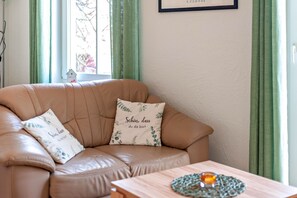 A tan leather sofa with two decorative pillows, a small house decoration on the windowsill, and a wooden table with a candle and a woven placemat. - Apartment 'Abendrot' with Wi-Fi (Ostseebad Kühlungsborn)