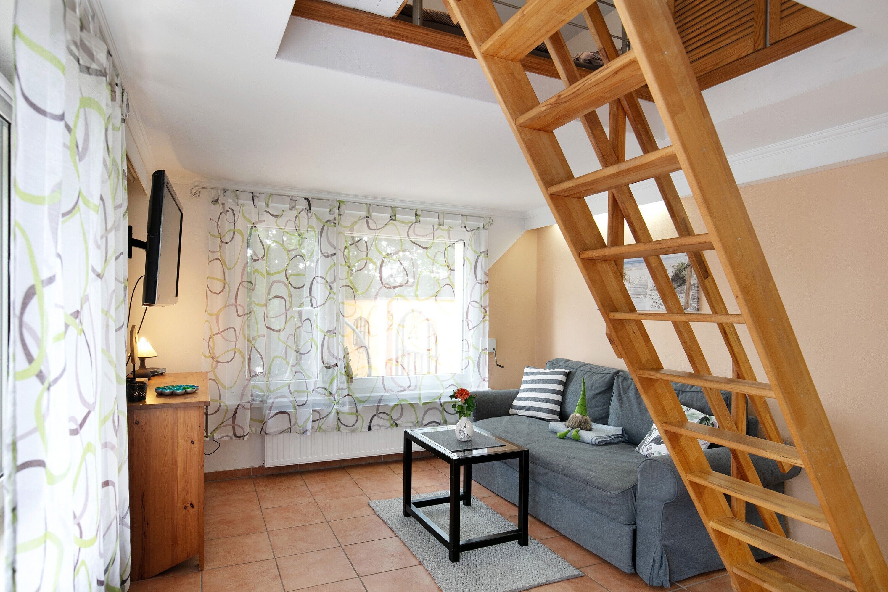 Un salon avec un canapé, une petite table, une échelle en bois et une fenêtre ornée de rideaux à motifs.