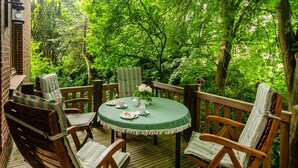 A wooden deck with a round table and chairs, surrounded by lush greenery.