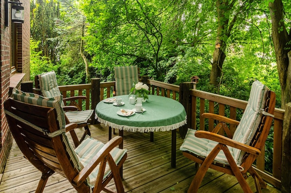 A wooden deck with a round table and chairs, surrounded by lush greenery.