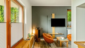 A modern living room with a flat-screen TV, wooden furniture, and a view of greenery outside.