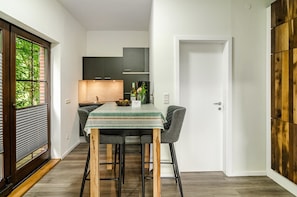 A modern kitchen with dark cabinets, a dining table with a green tablecloth, and a view of greenery through a window.