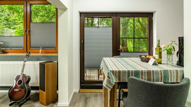 A room with a dining table, chairs, a guitar, speakers, and a view through a window with blinds.