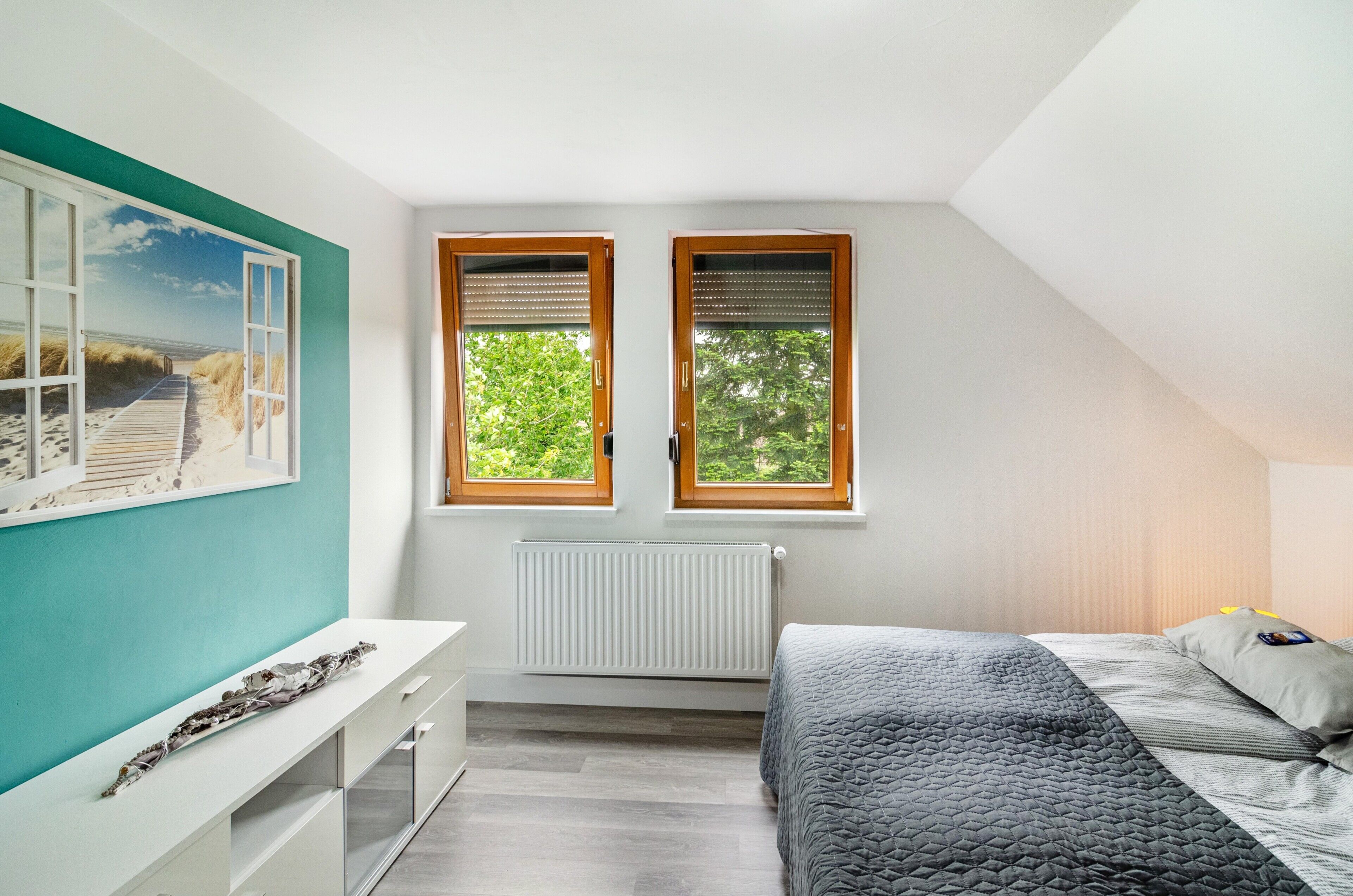 A bedroom with a bed, a window with a view of greenery, a radiator, and a white dresser.