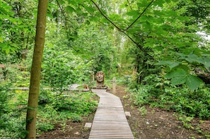 A wooden pathway in a forested area with dense greenery and a large stone sculpture.