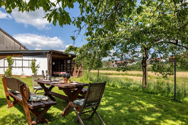 An outdoor dining area with a wooden table and chairs, surrounded by lush greenery and a view of a building in the background.