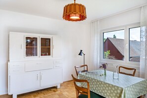 Et spiseområde med et bord, stole, et skab med glaslåger, en hængende lampe og et vindue med udsigt til en bygning.