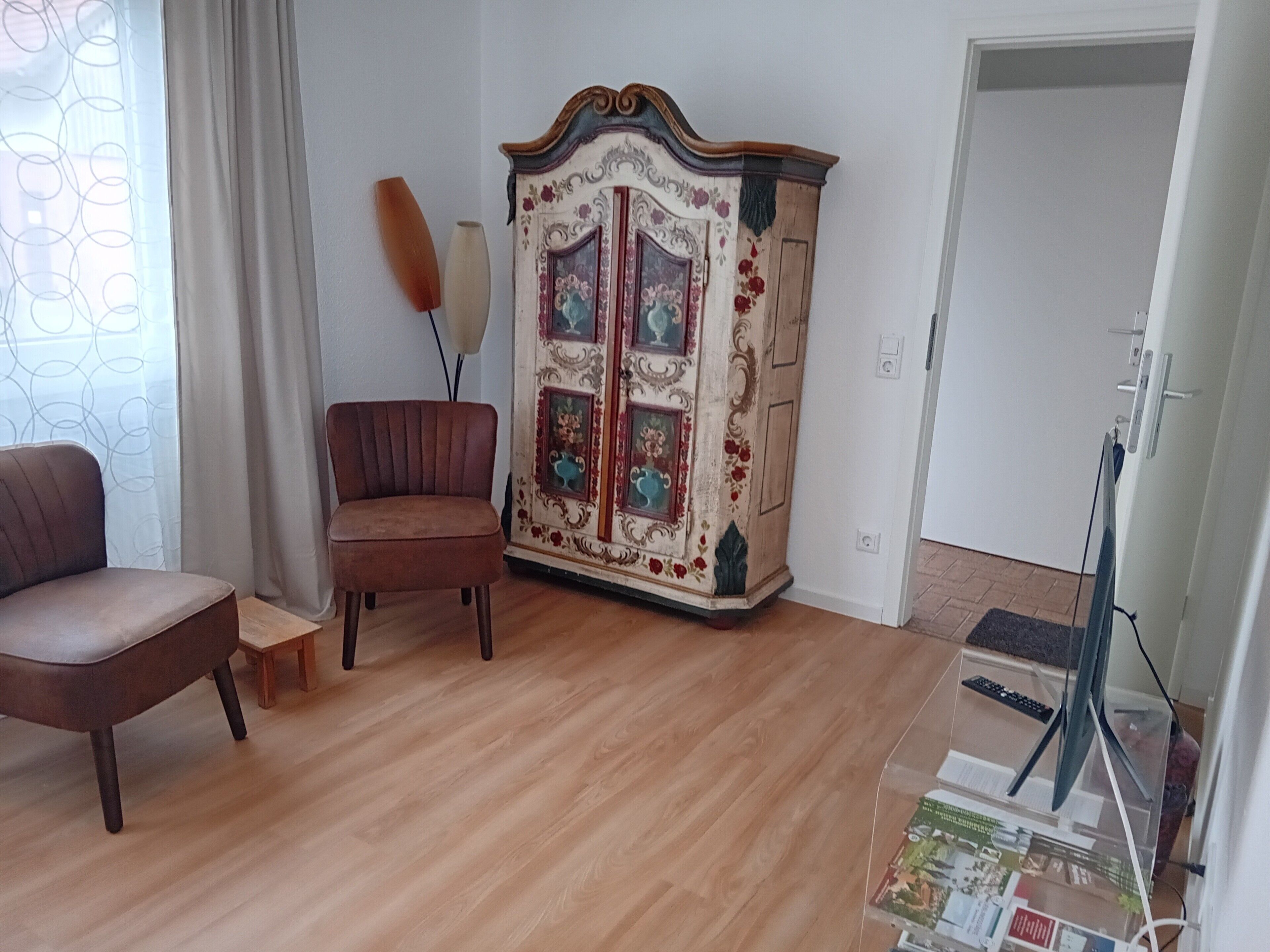 A room with a wooden floor, a brown armchair, a decorative cabinet with floral designs, and a small table.