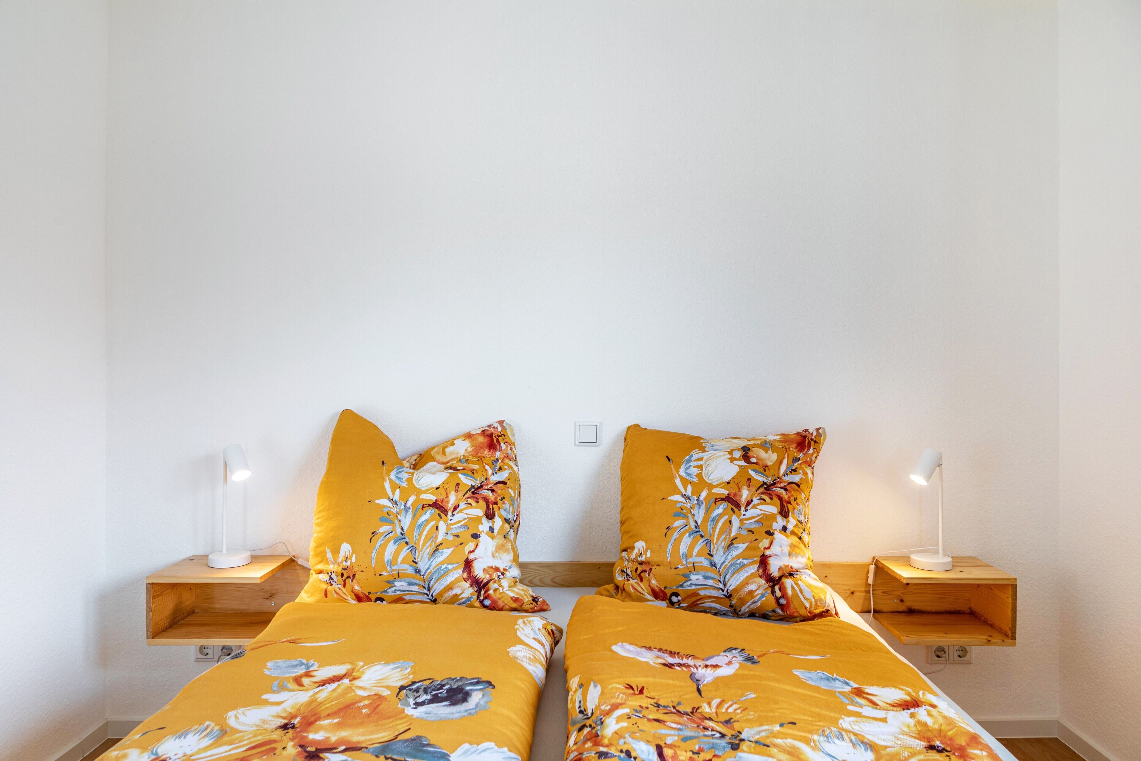 A room with two beds, each with a floral bedspread and matching pillows, flanked by bedside tables with lamps.
