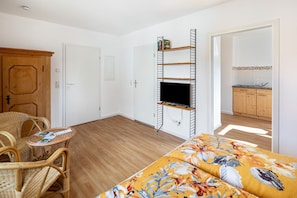 A room with a bed, a television mounted on a wall, wicker furniture, and a kitchen area visible through an open door. - Ferienhof im Salzbachtal ­ –  Wildbiene Plus für bis zu sechs Personen (Freiensteinau)