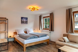 A bedroom with a wooden bed, a nightstand, a table, a sofa, a window with curtains, and a ceiling light fixture.