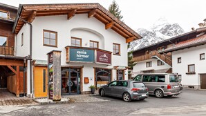 Un bâtiment portant une enseigne avec les noms « Xenia Hirmer » et « Berg Chalet », caractérisé par une façade en bois, une voiture garée devant et un paysage montagneux en arrière-plan.