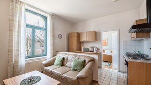 Ein kompakter Wohnbereich mit Küchenzeile, einer Couch mit Kissen, einem Esstisch und einem Fenster mit Vorhängen.