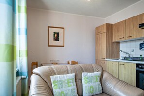 Un salon comprenant un canapé, deux coussins, un tableau encadré accroché au mur, une table à manger avec des chaises, et un coin cuisine avec des meubles en bois et un évier.