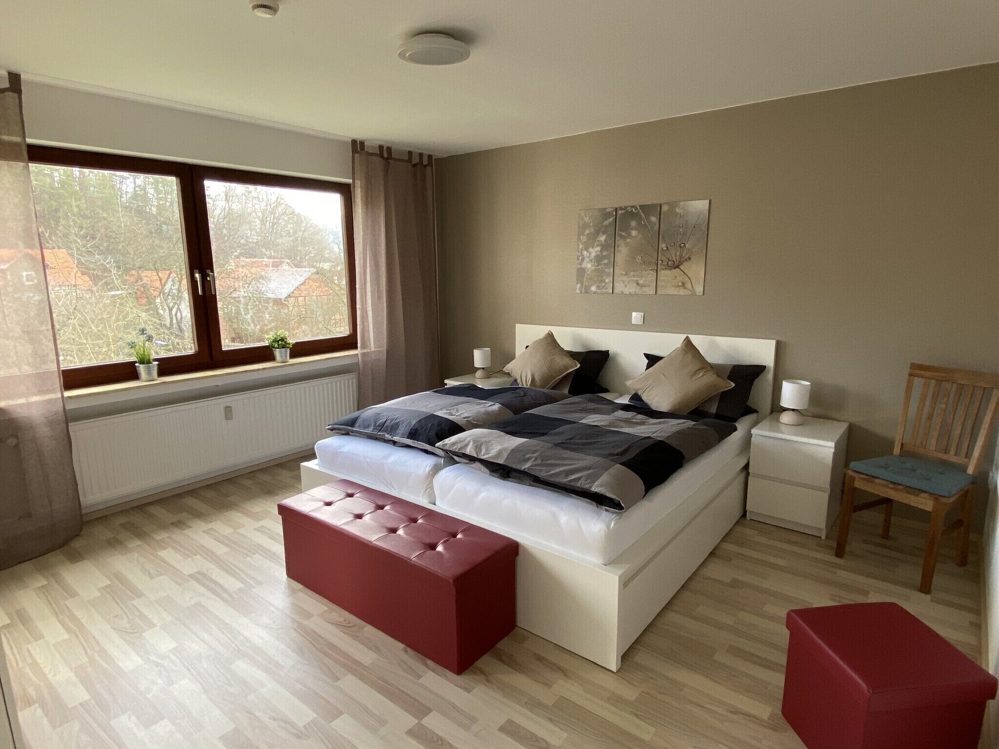 A bedroom with a bed, bedside tables, a chair, a red bench, a window with curtains, and a radiator.